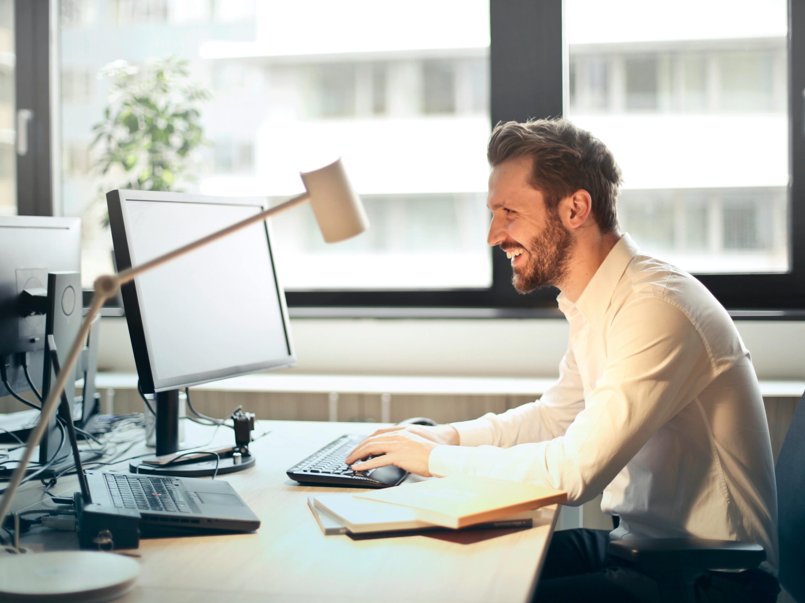 A man smiling while working at an office desk with a computer and natural daylight streaming in through large windows.